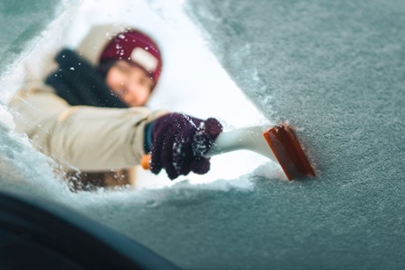 A woman cleans her windshield with an ice scraper.